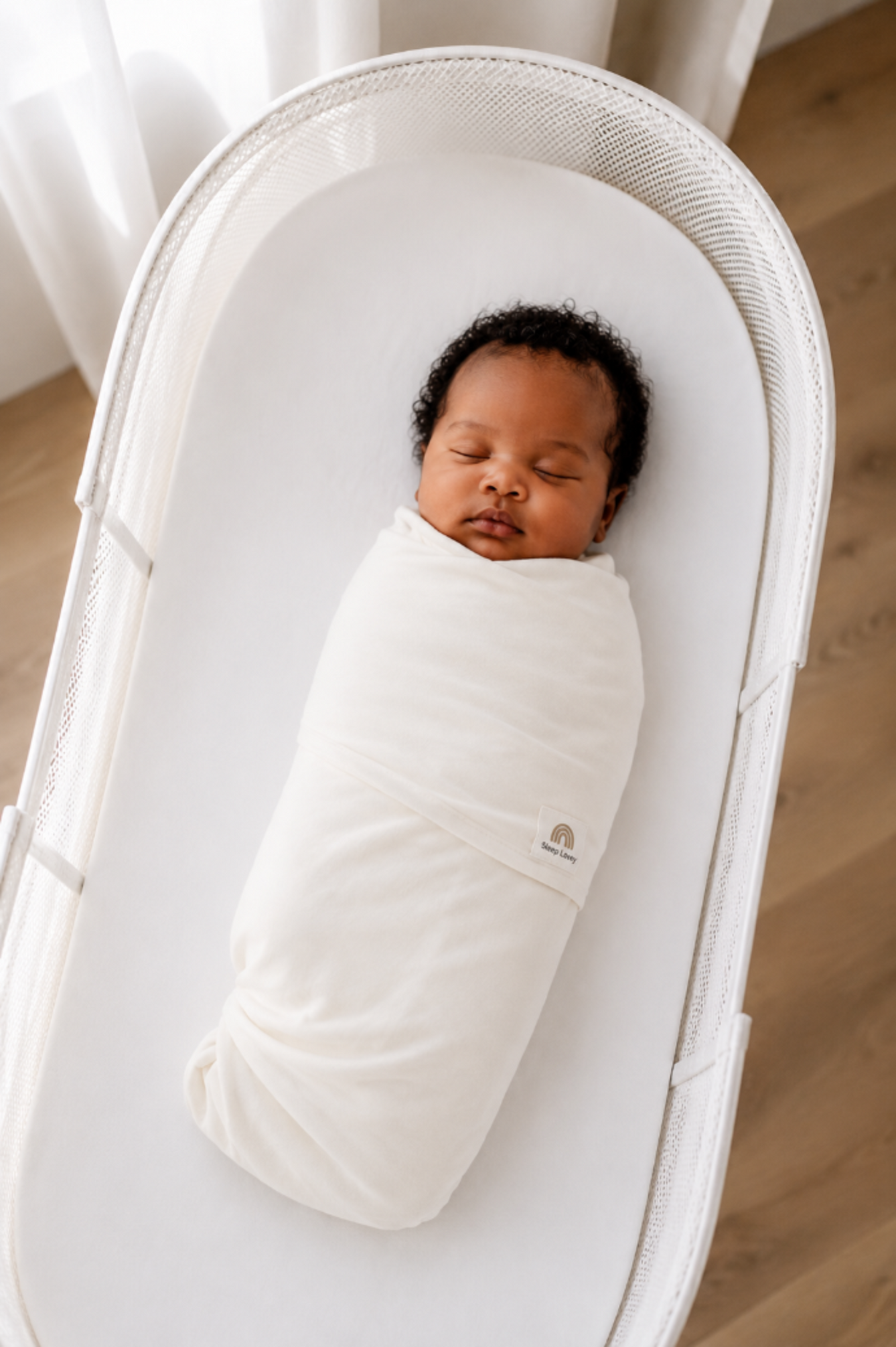 A sleeping newborn baby swaddled in a white swaddle blanket, lying inside a white snoo oval bassinet. 